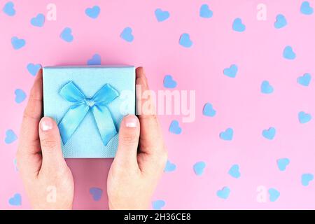 Geschenkschachtel aus Papier mit einer Schleife in den Händen der Frau auf einem rosa Hintergrund mit Konfetti-Herzen. Stockfoto
