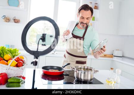 Foto eines professionellen Mannes aus dem lustigen Alter, der mit blauen T-Shirt-Schürze bekleidet ist, der im Wohnzimmer kochende Suppe kocht Stockfoto
