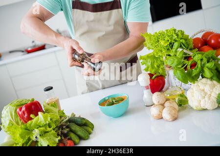 Zugeschnittenes Foto von Mann Hände würzen leckere Mahlzeit halten Pfefferbox tragen Küchengeschirr Schürze in Küche Wohnung drinnen Stockfoto