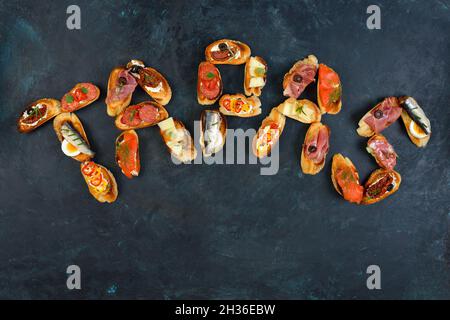 Köstliche gemischte spanische Tapas mit typischen Spezialitäten auf gebratenem Baguette mit Sesam in Form von Blockbuchstaben auf dunklem Hintergrund mit Kopierfläche Stockfoto