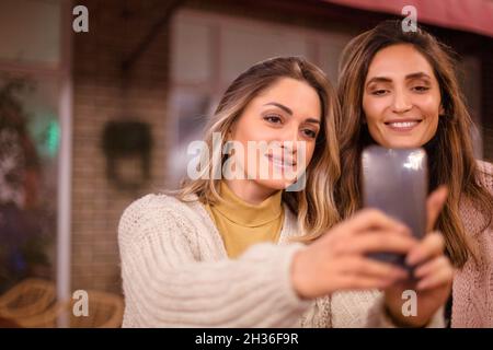 Zwei glückliche junge Frauen, weibliche beste Freundinnen, die Selfie auf dem Smartphone machen und beim Treffen am Wochenende im Café lächeln, fröhliche Freundinnen, die p Stockfoto