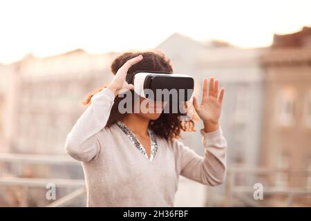 Technologie der Zukunft. Junge, moderne Mixed-Race-Frau in VR-Brille Headset erkunden virtuelle Realität im Freien mit Stadtbild auf dem Hintergrund, weibliche versuchen Stockfoto