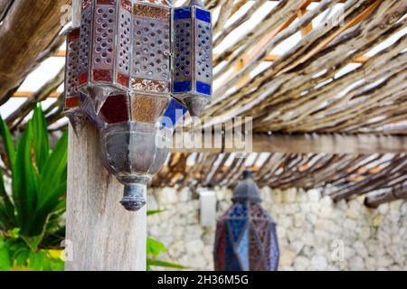 Nahaufnahme von antiken und exotischen dekorativen Eisenlaternen, die mit rotem, orangefarbenem und blauem Glas an einer Holzsäule und einem Holzdach hängen. Keine Personen. Stockfoto