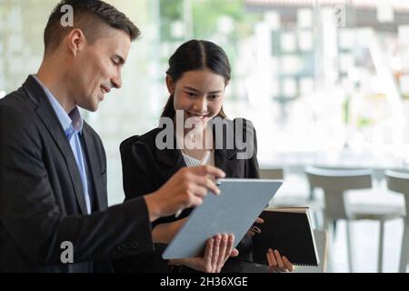 Fokussierte Mitarbeiter verschiedener ethnischer Herkunft führen Brainstorming auf einem digitalen Tablet durch, asiatische und kaukasische Kollegen tauschen Ideen am Schreibtisch aus Stockfoto