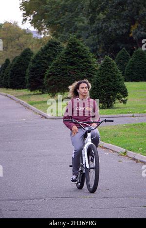 Ein junger Mann mit langen Haaren fährt mit dem Fahrradfahren, ohne sich am Lenker zu halten. In Flushing Meadows Corona Park in Queens, New York. Stockfoto