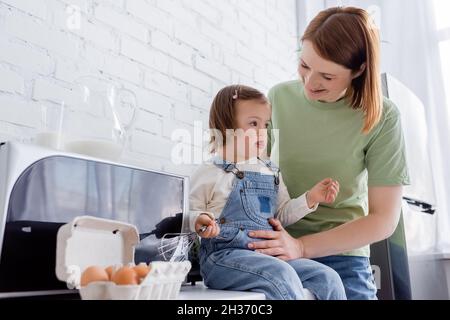 Lächelnde Mutter hält Tochter mit Down-Syndrom in der Nähe von Eiern und Milch in der Küche Stockfoto
