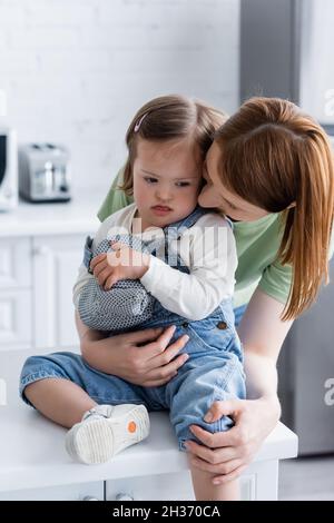 Mutter umarmte Tochter mit Down-Syndrom, die Zunge herausstreckt und in der Küche einen Backhandschuh hält Stockfoto