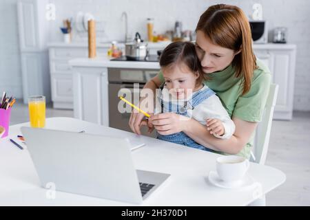 Mutter hält Farbstift in der Nähe des Kindes mit Down-Syndrom und Laptop in der Küche Stockfoto