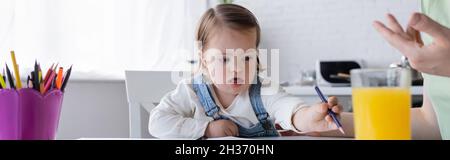 Kleinkind Mädchen mit Down-Syndrom hält Farbe Bleistift in der Nähe von Orangensaft und Mama, Banner Stockfoto