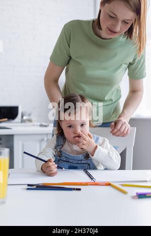 Kind mit Down-Syndrom mit Farbstift in der Nähe der Mutter und Skizzenbuch in der Küche Stockfoto