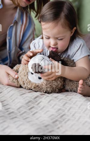 Baby-Mädchen mit Down-Syndrom hält weiches Spielzeug in der Nähe verschwommene Mutter auf dem Bett Stockfoto