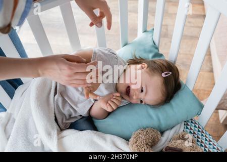 Draufsicht der Mutter, die die Hand eines fröhlichen Kindes mit Down-Syndrom in der Krippe hält Stockfoto