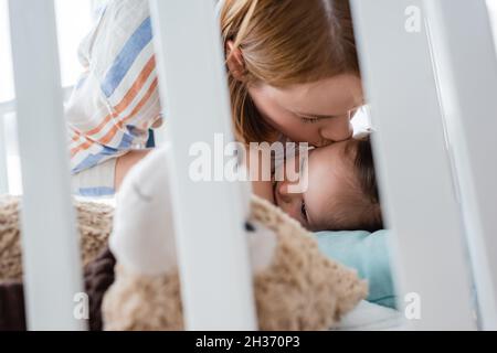 Mutter küsst Mädchen mit Down-Syndrom in der Nähe von weichen Spielzeug in Babykrippe Stockfoto