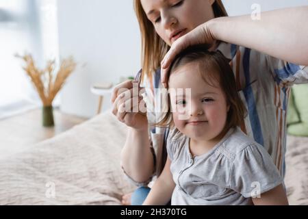 Verschwommene Mutter hält Haarspange in der Nähe der Tochter mit Down-Syndrom im Schlafzimmer Stockfoto