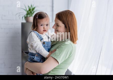 Lächelnde Mutter hält Kleinkind mit Down-Syndrom zu Hause Stockfoto