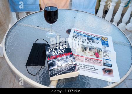 Reisekonzept nach COVID 19. Tisch vor einer spanischen Villa mit einer chirurgischen Gesichtsmaske, einem Glas Rotwein, Costa Blanca News und einem Buch mit dem Titel Lockdown Stockfoto