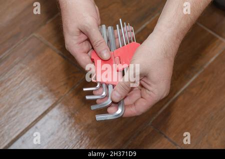 Hände halten einen Satz Sechskantschlüssel gegen den braunen Holzboden. Ein erwachsener Mann mit einem Handwerkzeug. Blick von oben aus einem Winkel. Selektiver Fokus. Stockfoto
