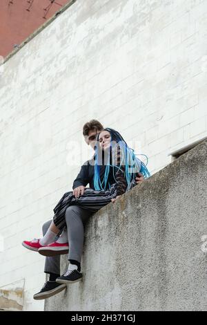 Ein Mann und eine Frau umarmen sich, sitzen auf einer Betonbrüstung. Die Frau ist lässig mit langen blauen Dreadlocks gekleidet. Mann in Geschäftskleidung. Blick vom Th Stockfoto