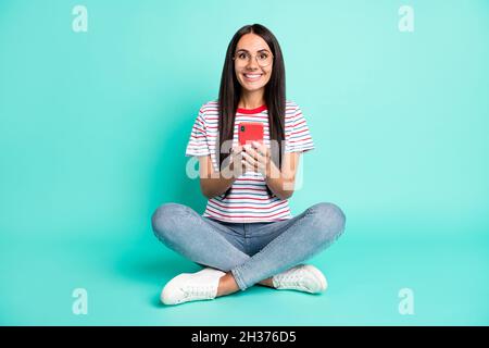 Foto in voller Größe von optimistischen Brünette Frisur Dame sitzen schreiben Telefon tragen T-Shirt Jeans Sneakers isoliert auf blauem Hintergrund Stockfoto