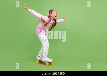 In voller Länge Foto von beeindruckt kleine blonde Mädchen Ride Skate tragen Kapuzenhose Sneakers isoliert auf grünem Hintergrund Stockfoto