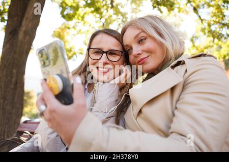 Low-Angle-Aufnahme von reifen Freundinnen, die Spaß haben, mit Smartphone im Park Stockfoto