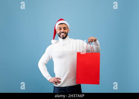 Glücklicher arabischer Mann mit Weihnachtsmütze und Einkaufstasche mit Weihnachtsgeschenk auf blauem Hintergrund, Studioaufnahme Stockfoto
