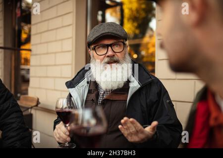 Das verkürzte Porträt eines stilvollen älteren Mannes, der sich am warmen Herbsttag mit Freunden im Café traf und Rotwein trank Stockfoto