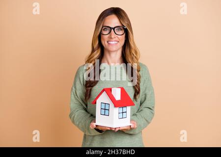 Foto Porträt Frau trägt Brille lächelnd halten kleinen Haus isoliert pastellbeige Farbe Hintergrund Stockfoto