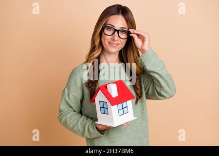Foto Porträt weibliche Agent trägt Brille lächelnd halten kleine Haus neu zu Hause isoliert pastellbeige Farbe Hintergrund Stockfoto