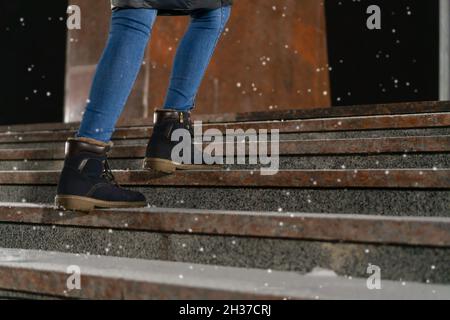 Frauenfüße in blauen Stiefeln erklimmen an einem frostigen Abend im Licht von Straßenlampen die Treppe mit etwas Schnee, um Platz zu kopieren Stockfoto