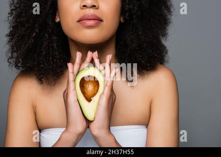 Beschnittene Ansicht einer afroamerikanischen Frau in weiß oben, die eine Hälfte der Avocado isoliert auf grau hält Stockfoto