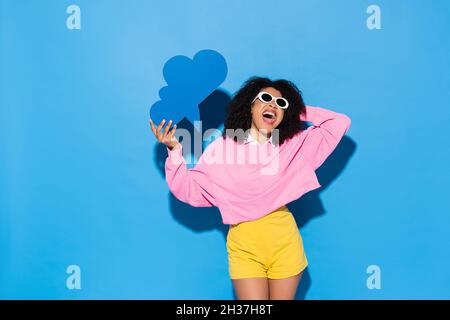 Erstaunt afroamerikanische Frau in Sonnenbrille mit leeren Gedanken Blase und Schreien auf blau Stockfoto