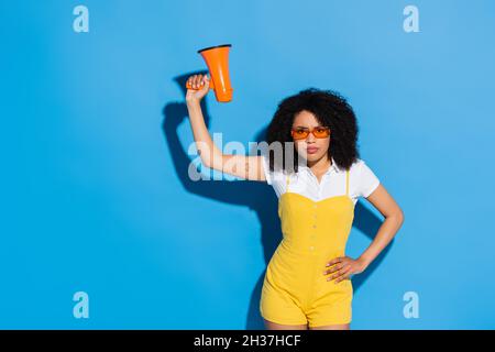 Unzufrieden african american Frau mit Hand auf Hüfte hält Megaphon auf blau Stockfoto