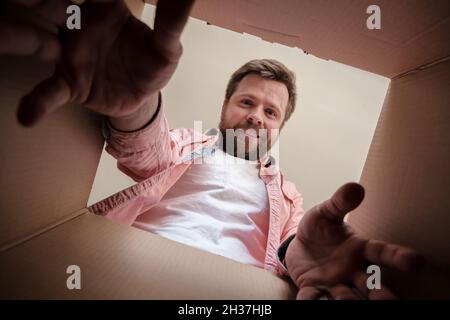 Ein zufriedener lächelnder Mann packt eine gelieferte Schachtel mit einem Paket oder einem Geschenk aus, greift nach draußen und nimmt die Bestellung auf. Auspacken der Innenansicht. Stockfoto