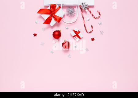 Weihnachten, Neujahr. Weiße Papiertüte mit Geschenken, süßen Bonbons, Weihnachtsdekorationen, roten Kugeln, silbernen Glitzer-Konfetti-Sternen auf pastellrosa Hintergrund. Flach liegend, Kopierplatz Stockfoto