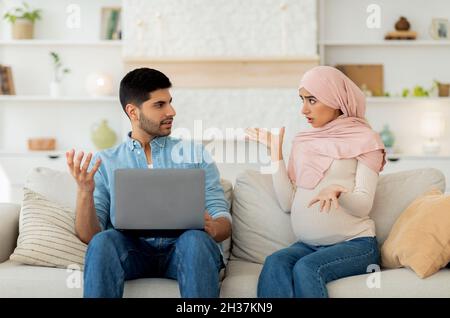 Gestresste, schwanger-muslimische Frau, die ihren Mann anschrie, während der Mann einen Laptop benutzte und zu Hause auf dem Sofa saß Stockfoto