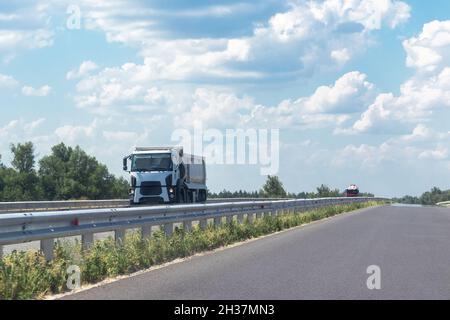 Ein weißer Kipper fährt an einem sonnigen Tag auf einer flachen Asphaltstraße zwischen Feldern vor einem blauen Himmel mit Wolken aus der Stadt. Eine Maschine für den Transport von BU Stockfoto