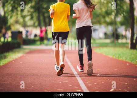 Nahaufnahme der Beine in Turnschuhen von zwei Kindern auf der Laufstrecke des Stadions, Rückansicht. Junge und Mädchen auf der roten Joggingstrecke im Park laufen. Schulkinder Stockfoto