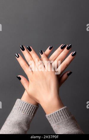 Weibliche Hände in grauem Strickpullover mit schöner Maniküre - dunkelschwarze Nägel auf grauem Hintergrund. Nagelpflegekonzept Stockfoto