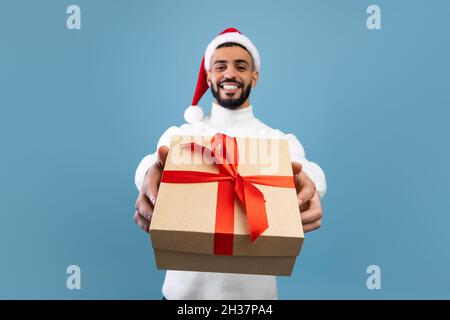 Neujahrsfest. Glücklicher araber in Weihnachtsmütze hält Weihnachtsgeschenk und dehnt es zur Kamera Stockfoto