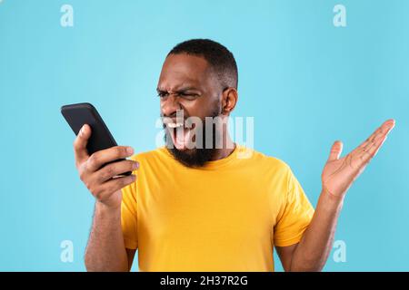 Wütend jungen schwarzen Kerl schreiend auf Smartphone über blauen Studio-Hintergrund. Stressmanagement und negative Emotionen Stockfoto