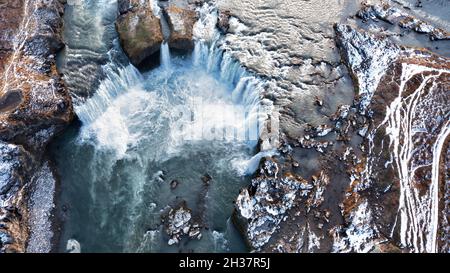 Drohnenaufnahme des Godafoss Wasserfalls, Island, aufgenommen von direkt oben. Luftaufnahme der mächtigen Kaskade, des Flusses und der schneebedeckten Felsen. Spätherbst, Stockfoto