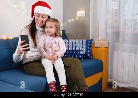 Familienweihnachtsfeier zu Hause während einer covid Pandemie, Online-Grüße, Anruf von Freunden und Verwandten per Smartphone. Mutter und Kind im Hut Stockfoto