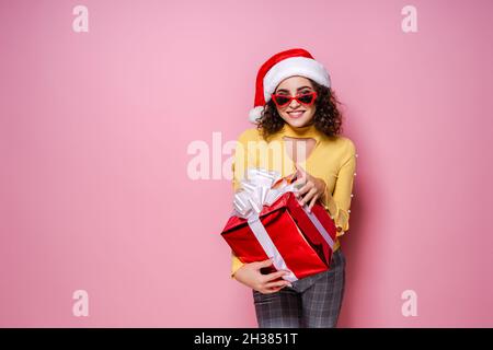 Fröhliches lockiges Mädchen in dem Hut des Weihnachtsmannes hält eine Sonnenbrille die geschenkbox, während sie auf einem rosa Hintergrund steht. Neujahr Stockfoto