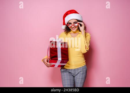 Glücklich lockiges Mädchen in Santa Hut, hält Brille präsent, während steht auf rosa Hintergrund. Neujahr Stockfoto