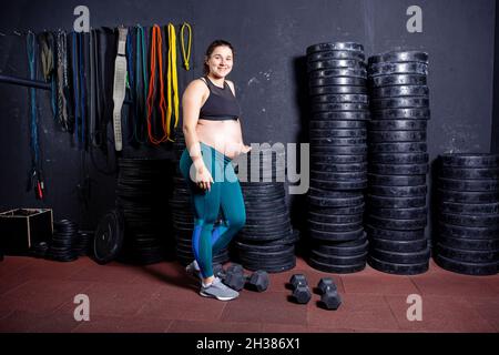 Starke Schwangerin posiert mit Kurzhanteln in den Händen vor dem Hintergrund von Sportgeräten im Fitnessstudio. Workouts und Übungen für Schwangeren in Stockfoto