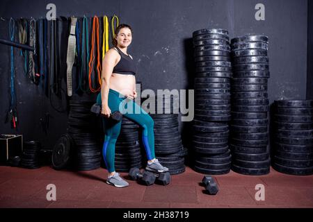 Starke Schwangerin posiert mit Kurzhanteln in den Händen vor dem Hintergrund von Sportgeräten im Fitnessstudio. Workouts und Übungen für Schwangeren in Stockfoto