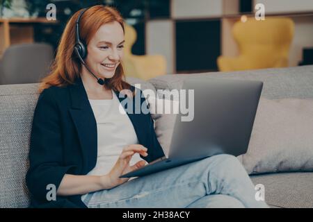 Junge positive Gingerhead-Studentin, die im Café ein Online-Webinar über Laptop und Headset führt Stockfoto