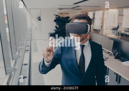 Junger Geschäftsmann in elegantem Anzug mit VR-Headset, berührt die Erforschung der virtuellen Realität bei der Arbeit Stockfoto