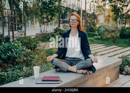 Entspannte Rotschopf junge Frau macht Yoga draußen sitzt in Lotusposen im Park Stockfoto